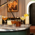 Five assorted cocktails on a marble bar counter in a lounge with paintings and a staircase in the background.