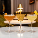 Three elegant cocktails on a table in a cozy bar setting with warm lighting.