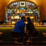 A couple shares a kiss at a bar counter, framed by an arch with a decorative background and bottles.